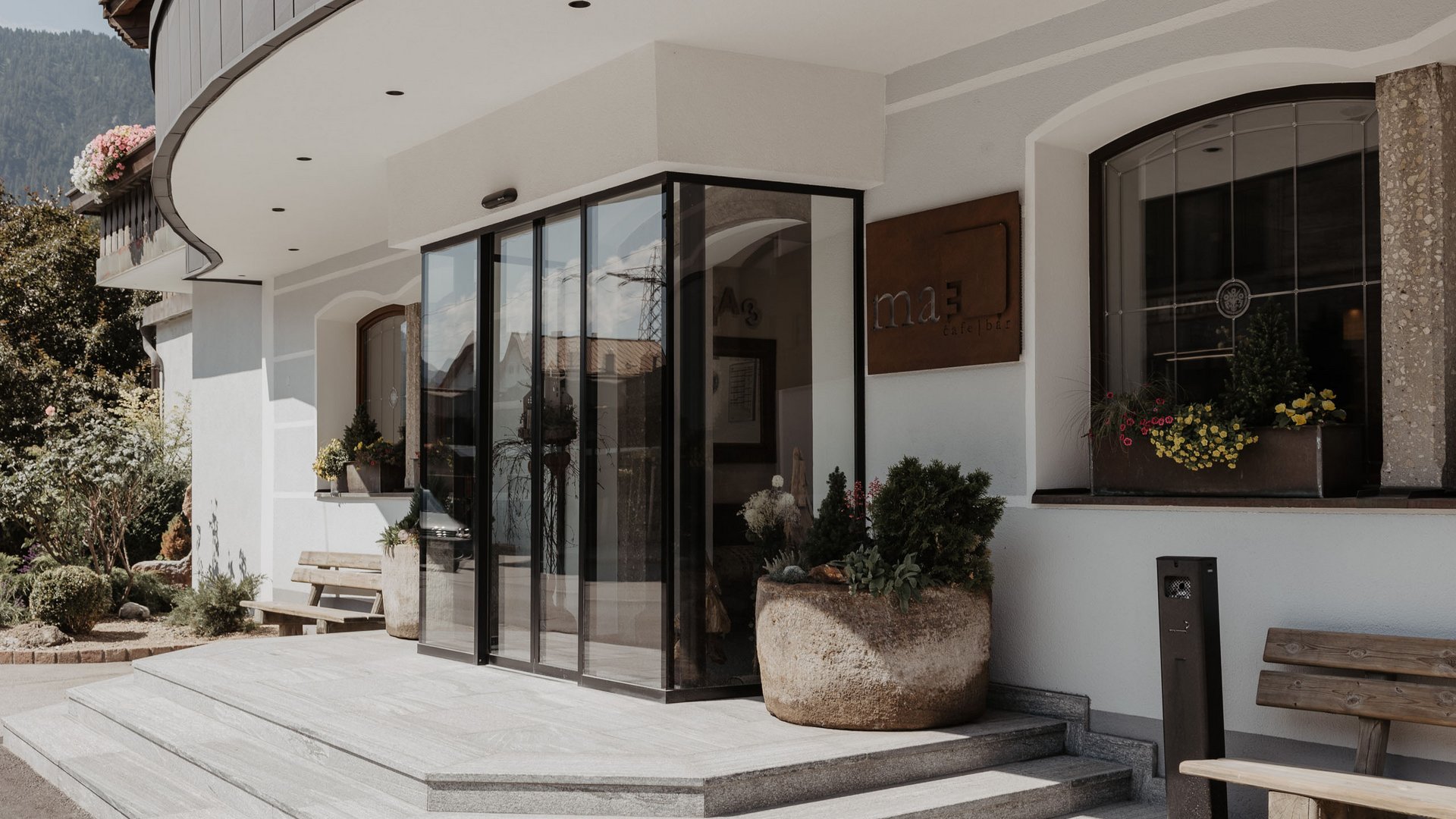 Dein Parkhotel in Matrei am Brenner Eingang mit Glasfront und Treppenstufen zu einem Café mit Blumen und Bänken